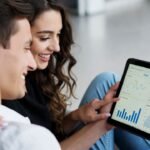 Casal jovem sorrindo e conversando enquanto analisam um tablet com gráficos de orçamento pessoal